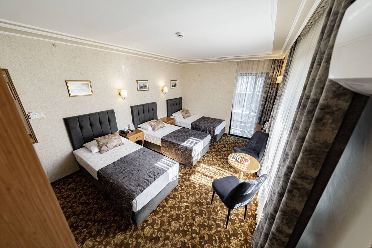 A hotel room with two beds, a small table, a chair, and a view of the city through the window.