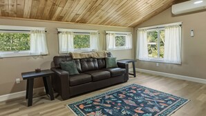 A room with a wooden ceiling, a brown leather sofa, a patterned rug, and three windows with curtains.