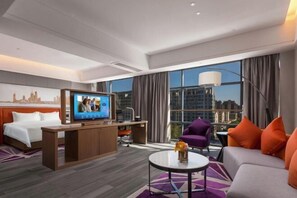 A hotel room with a bed, a desk, a TV, a sofa, and a balcony with a view of the city.
