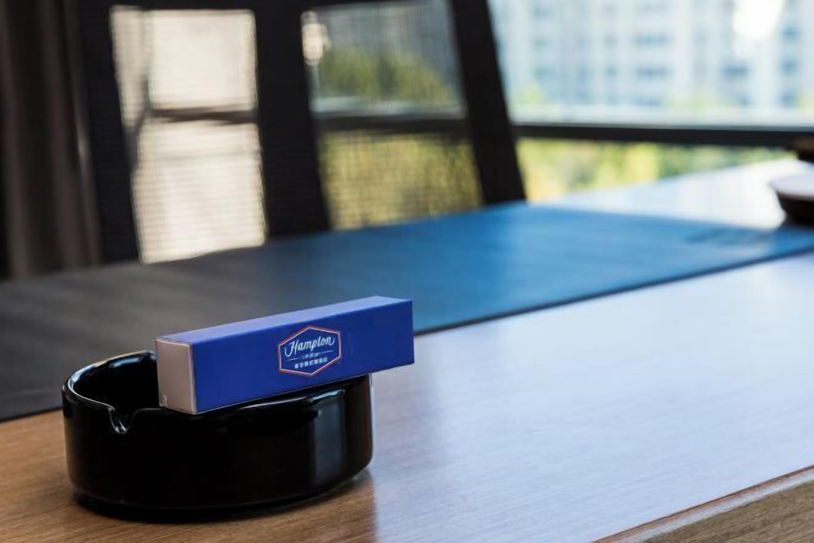 Un cendrier de la marque Hampton, posé sur une table en bois dans une pièce d’où l’on peut voir des bâtiments à travers une fenêtre.