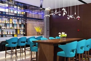 Zona de bar moderna con barra de madera y sillas azules. Se observan estantes con botellas y una lámpara decorativa colgante.