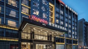 Moderni hotelli, jossa on suuri ja näkyvästi esillä oleva «Hampton»-kyltti, suuret ikkunat ja hyvin valaistu sisäänkäynti.