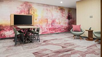 A modern room with a large abstract painting on the wall, a flat-screen TV mounted on a wooden stand, a small table with chairs, and a patterned carpet.
