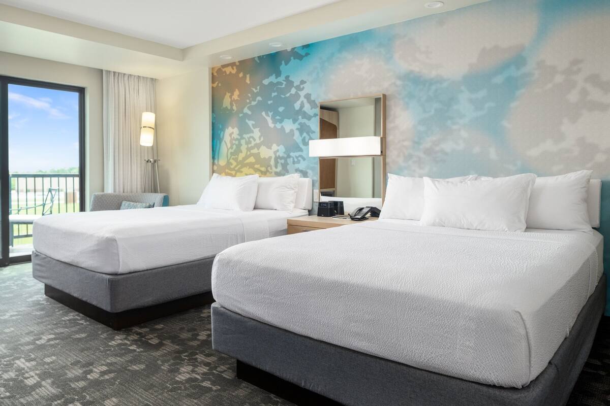A hotel room with two beds, a large window with a view, a mirror, and a lamp.