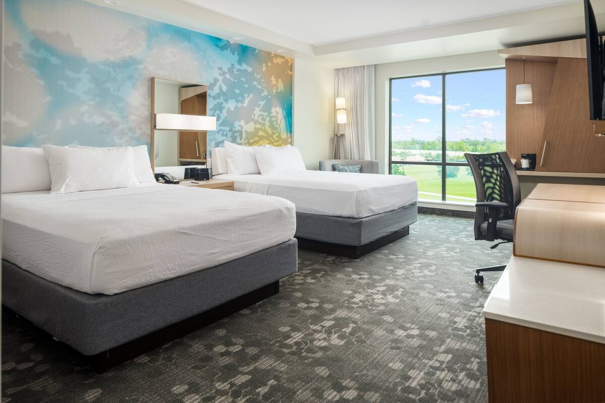 A hotel room with two beds, a desk, and a large window offering a view of the outdoors.
