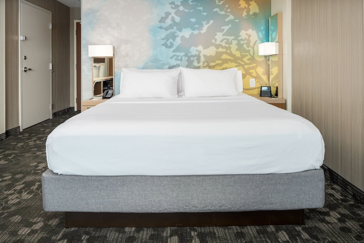 A hotel room with a large bed, a nightstand with a lamp, a mirror, and a wall with a camouflage pattern.