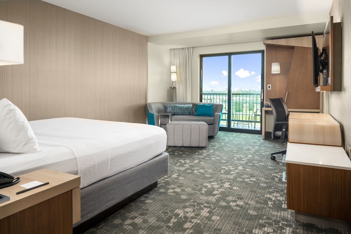 A hotel room with a large bed, a desk, a sofa, and a balcony with a view.
