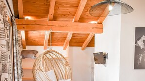 Ein gemütliches Zimmer mit Holzbalkendecke, einem Hängestuhl, einem Bett mit zusammengelegtem Handtuch, einem kleinen Tisch mit Laterne und gemusterten Vorhängen.