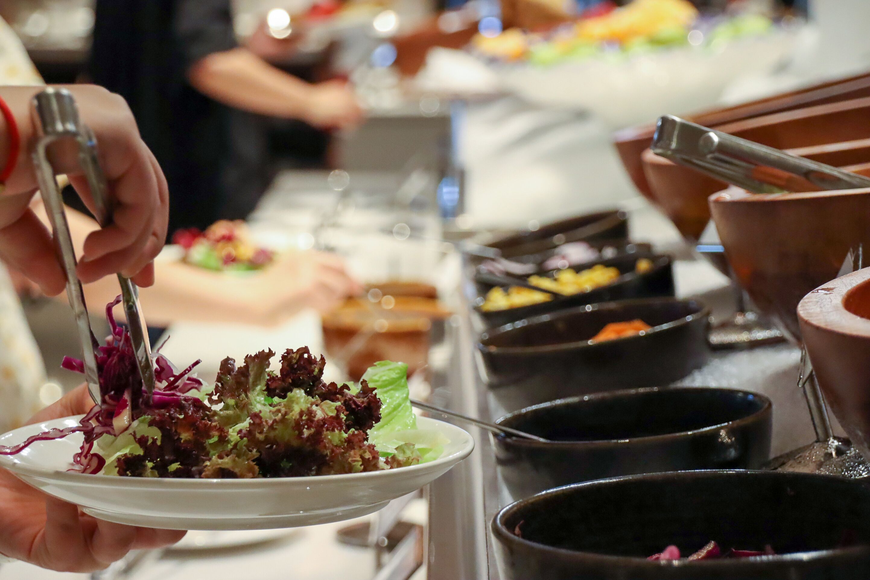A buffet setup with various food items in black bowls and a person using tongs to serve salad.