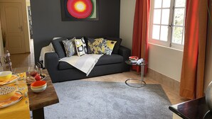 Ein modernes Wohnzimmer mit einem dunklen Sofa, bunten Kissen, einem Glastisch und einem Bild an der Wand.