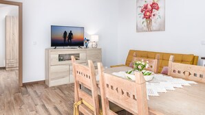 A modern living room with a wooden dining table, chairs, a TV, a sofa, and a painting on the wall.