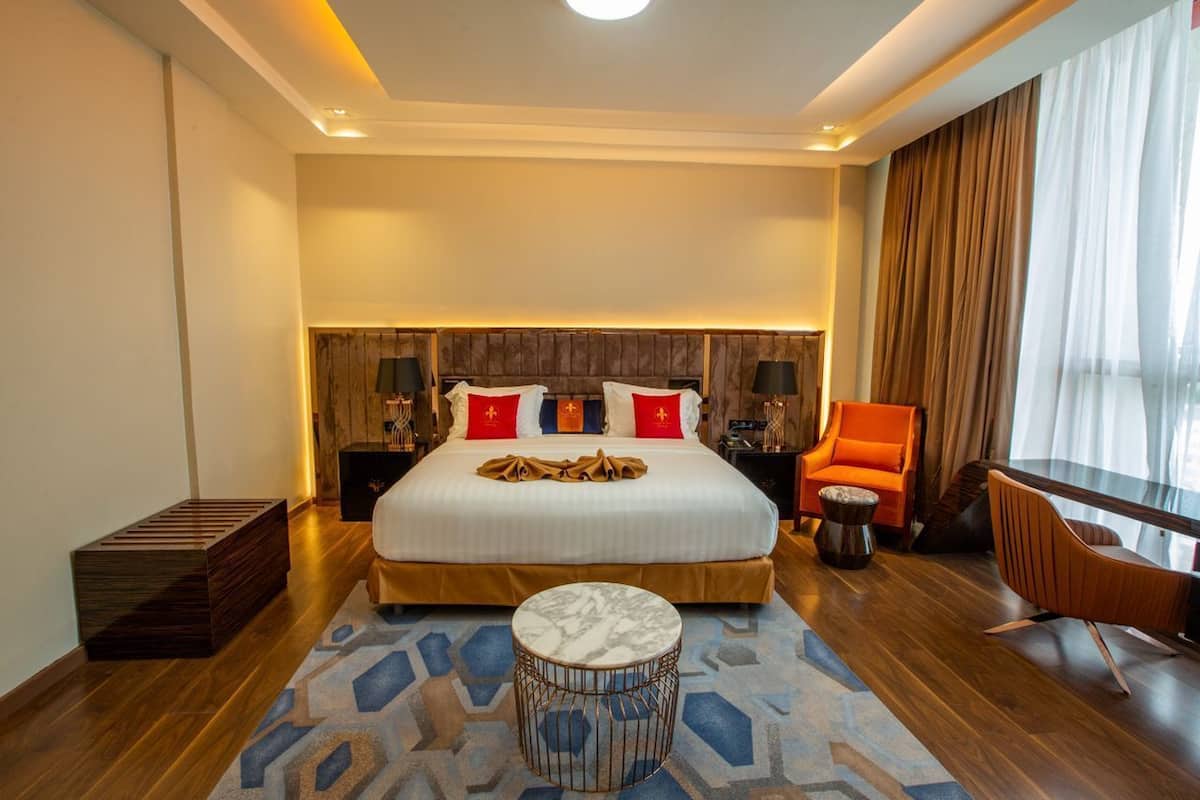 Habitación de hotel moderna con una cama grande, dos mesitas de noche con lámparas, una mesa de centro redonda, un banco de madera y vistas a la ciudad a través de amplios ventanales.