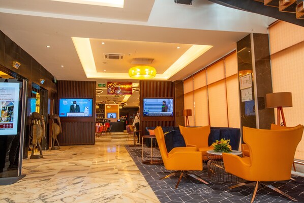Un vestíbulo de hotel con una recepción, zona de estar con sillas anaranjadas, una pantalla de televisión y una lámpara de araña grande.