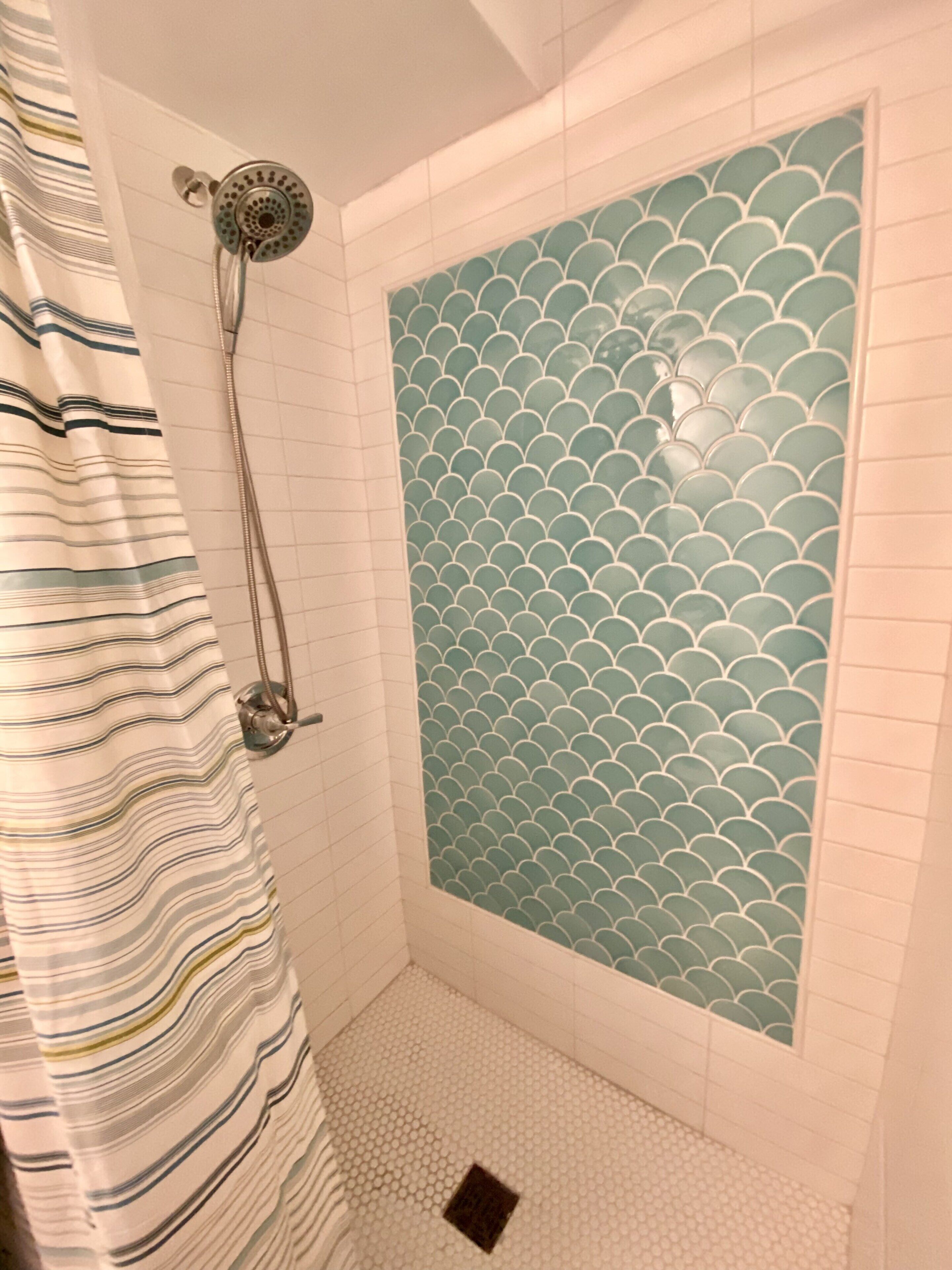 A shower with a scalloped tile design, a striped shower curtain, and a handheld showerhead.