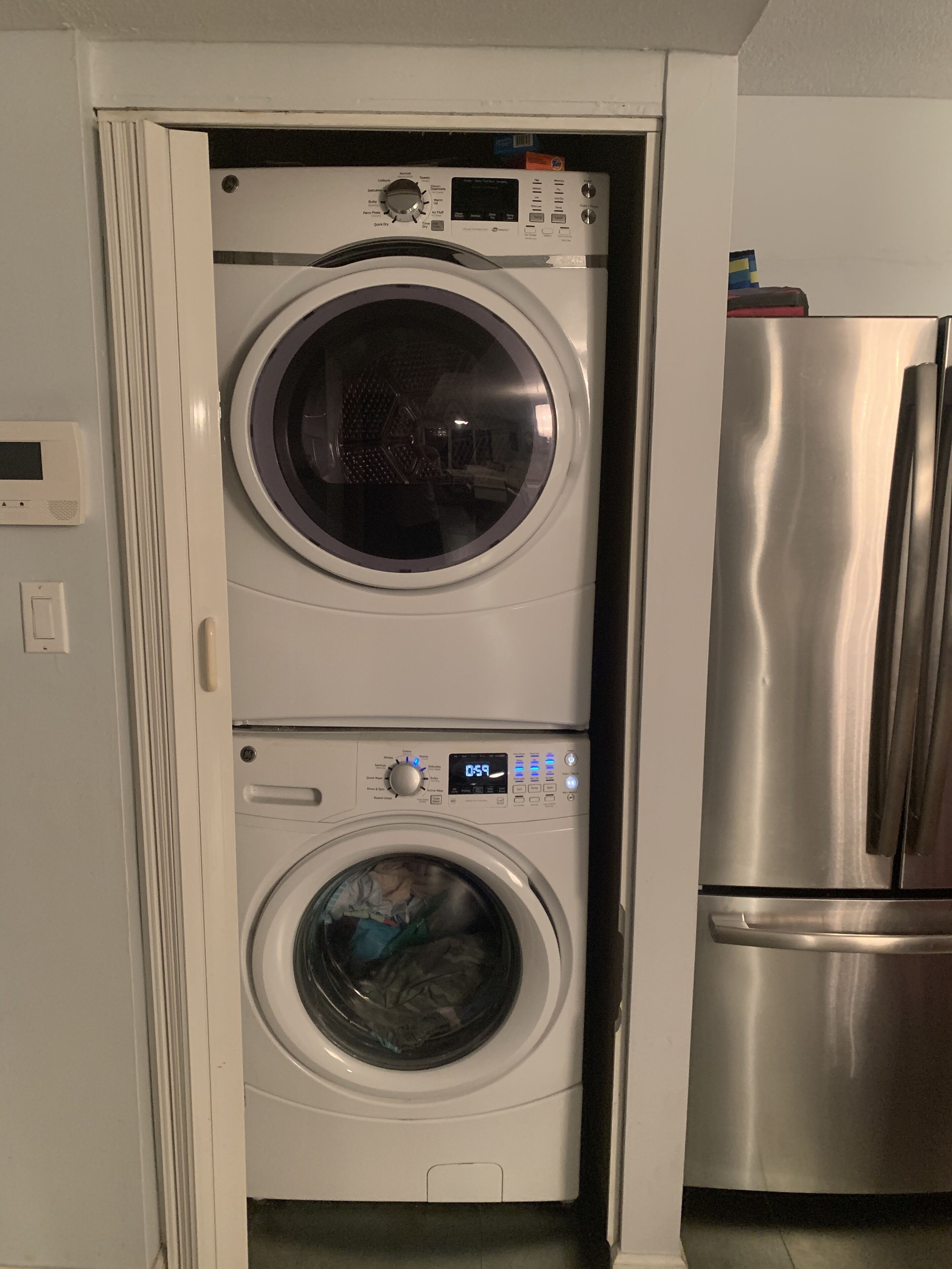 A compact washer-dryer unit with a digital display, a stainless steel refrigerator, and a white door frame.