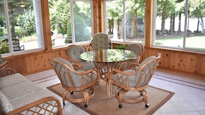 Une pièce aux murs lambrés, avec une table en verre entourée de chaises en osier, de grandes fenêtres et une vue sur les arbres.