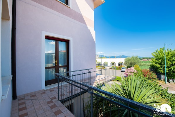 A balcony with a metal railing, a door with glass panels, and a view of a road and greenery.