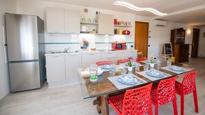 A modern kitchen with white cabinets, a stainless steel refrigerator, a microwave, and a dining area with a glass table and red chairs. - Il Cortiletto - Appartamento Lores (Manerba del Garda)