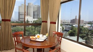 A dining area with a round table, two chairs, and a view of a cityscape through large windows. - Beach Luxury Hotel (Karachi)