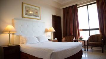Habitación de hotel con una cama grande, dos lámparas de noche, un cuadro enmarcado sobre la cama, una ventana con cortinas y dos sillas.