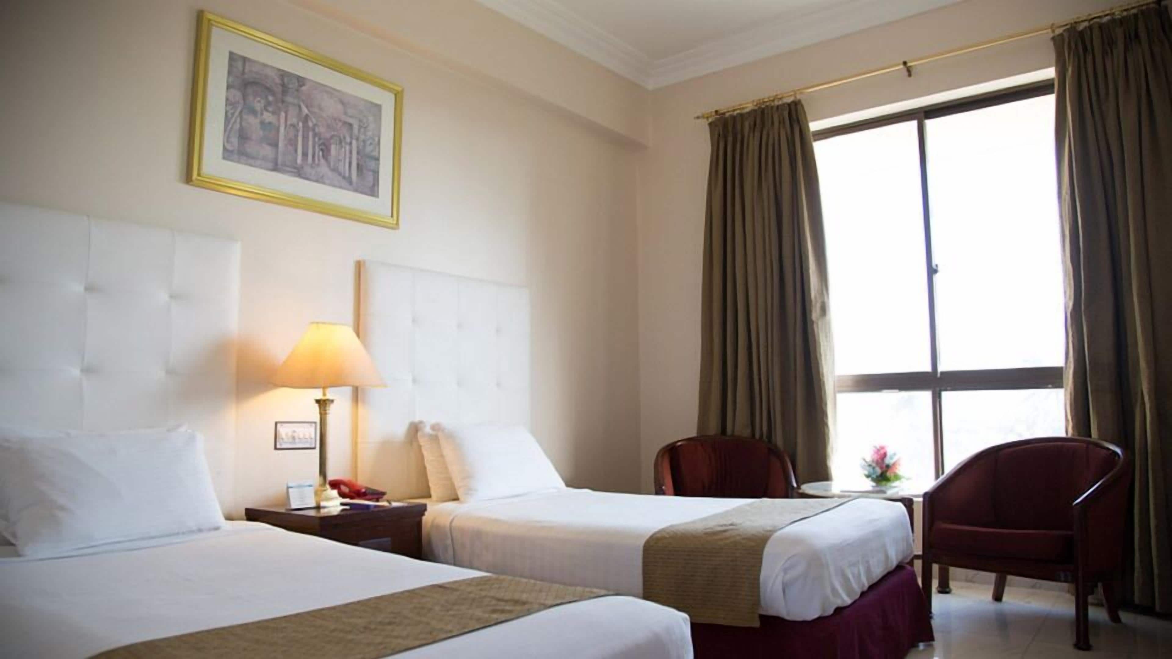 A hotel room with two beds, a nightstand, a lamp, a chair, and a large window with curtains.
