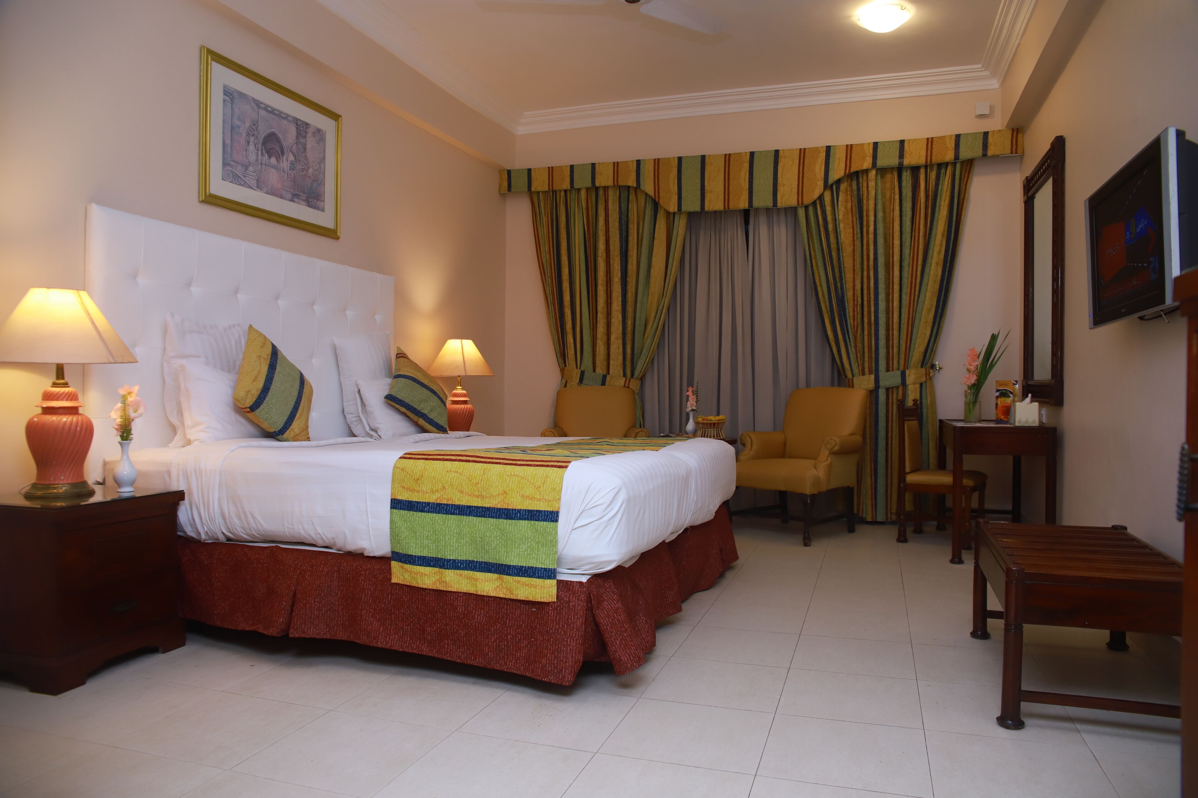 A hotel room with a large bed, two bedside tables with lamps, a chair, a wooden bench, and a television on the wall.