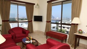 A spacious living room with red furniture, large windows with curtains, a flat-screen TV, and a view of the cityscape. - Beach Luxury Hotel (Karachi)