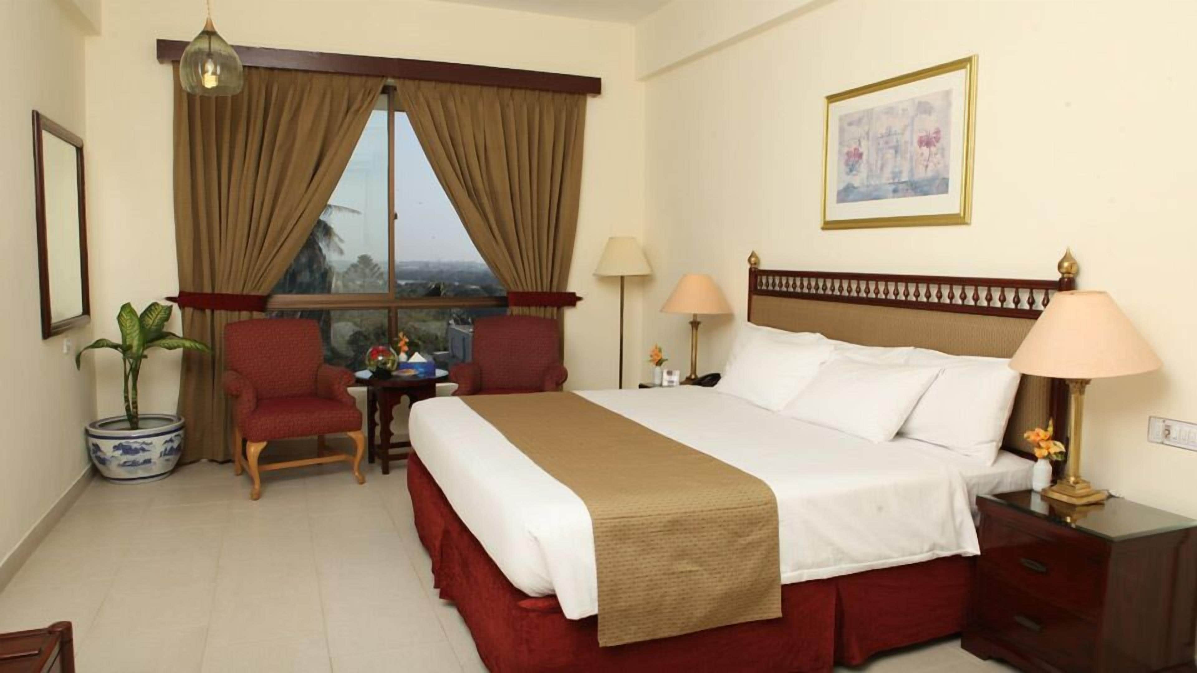 Quarto de hotel com uma cama grande, duas cadeiras, uma mesinha, um vaso de planta, uma janela com cortinas e um quadro na parede.