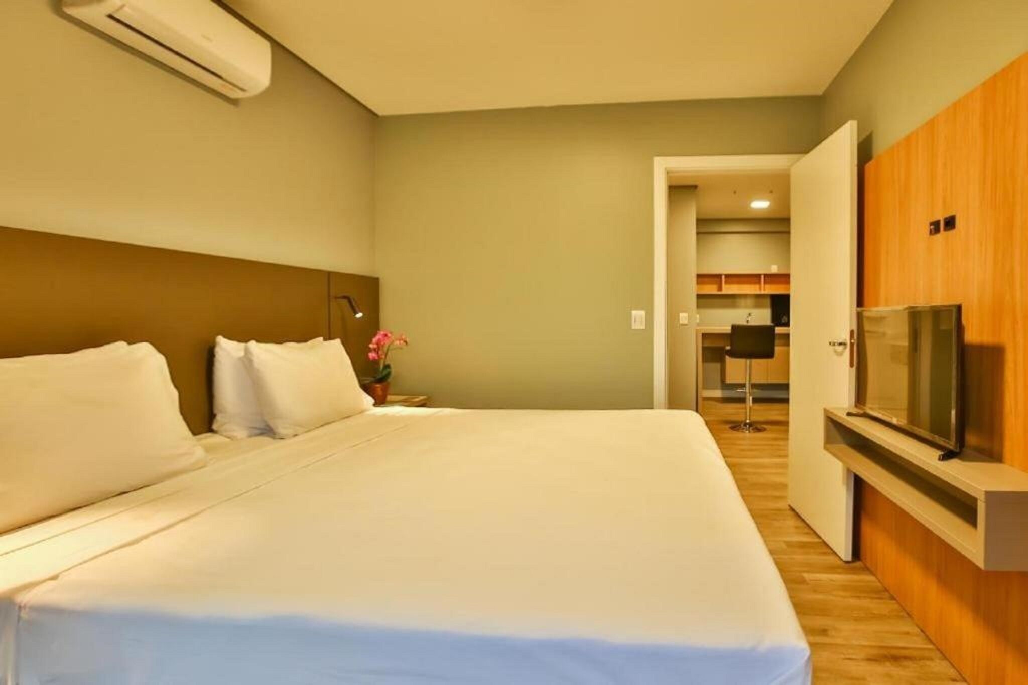 Quarto de hotel com uma cama grande, televisão, guarda-roupa de madeira e uma kitchenette ao fundo.