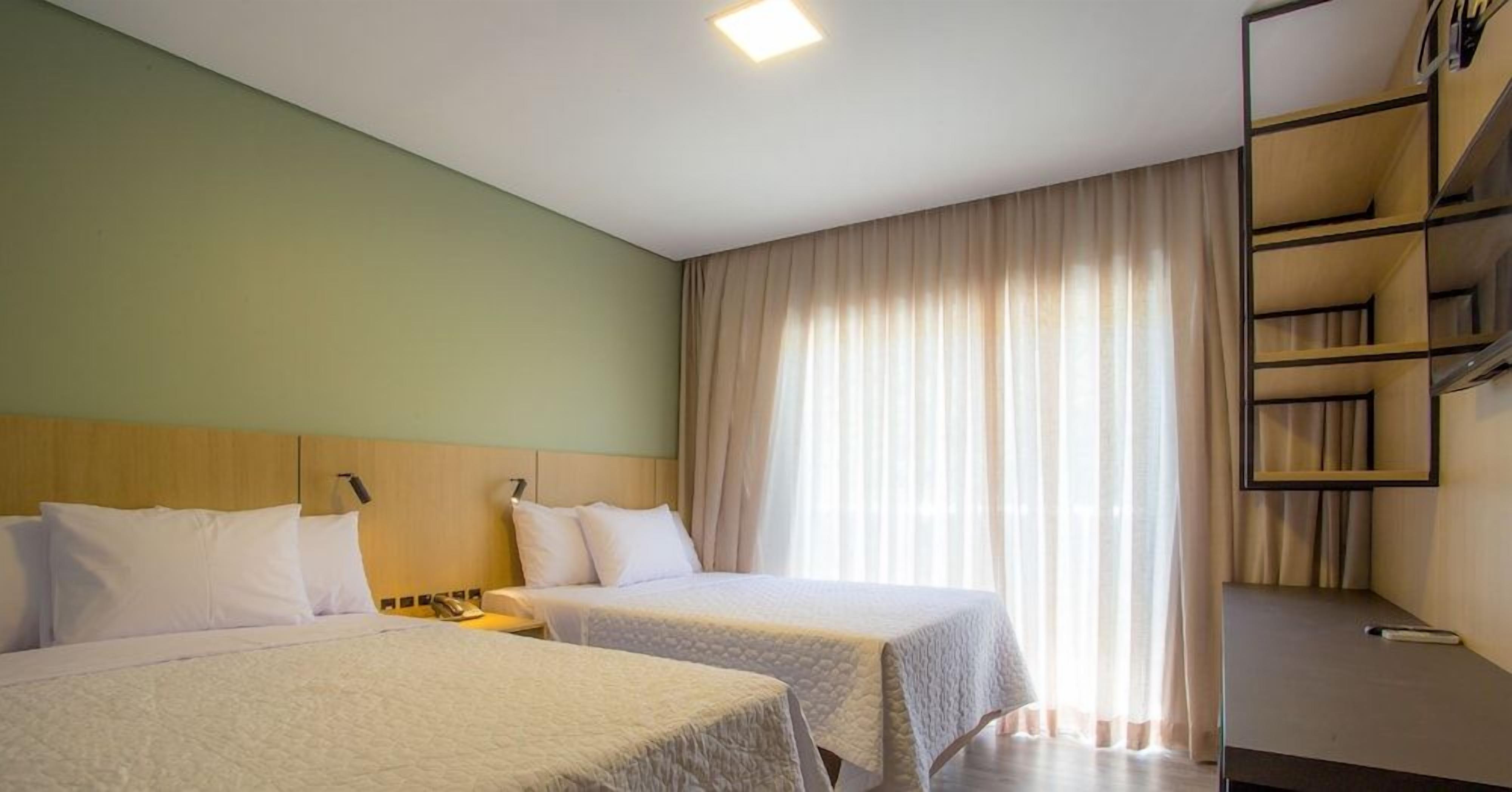 Quarto de hotel com duas camas, uma escrivaninha, uma cadeira, um guarda-roupa e uma janela com cortinas.