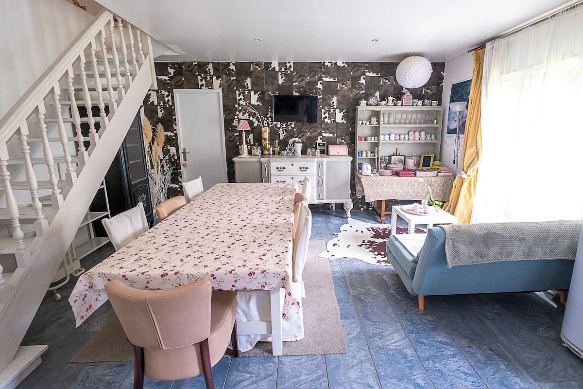 Un coin repas avec une table dressée pour deux personnes, un canapé, un escalier et un espace cuisine équipé d’un téléviseur.