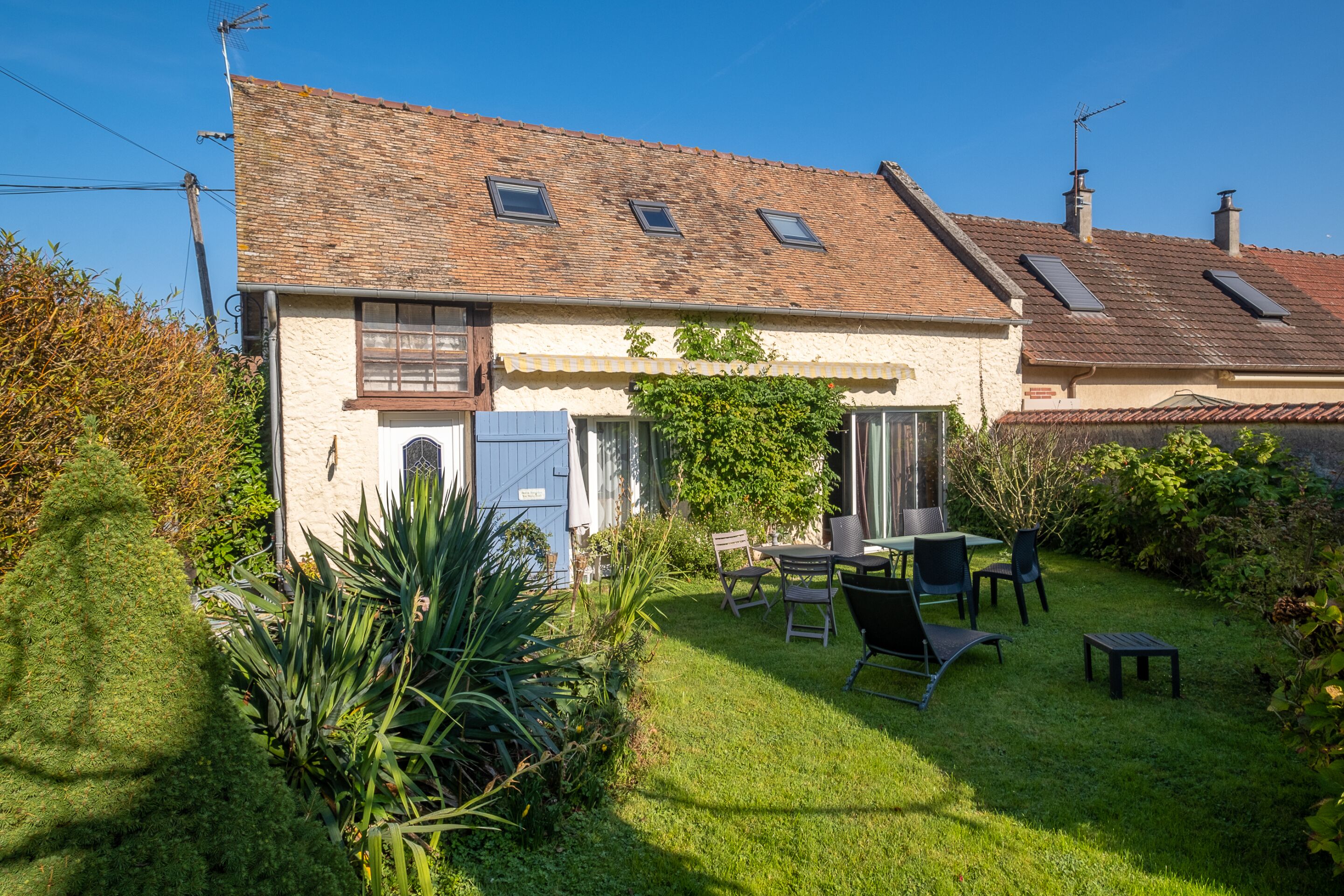 Une maison avec un toit en tuiles, un petit jardin avec une table et des chaises, et une fenêtre avec un volet.