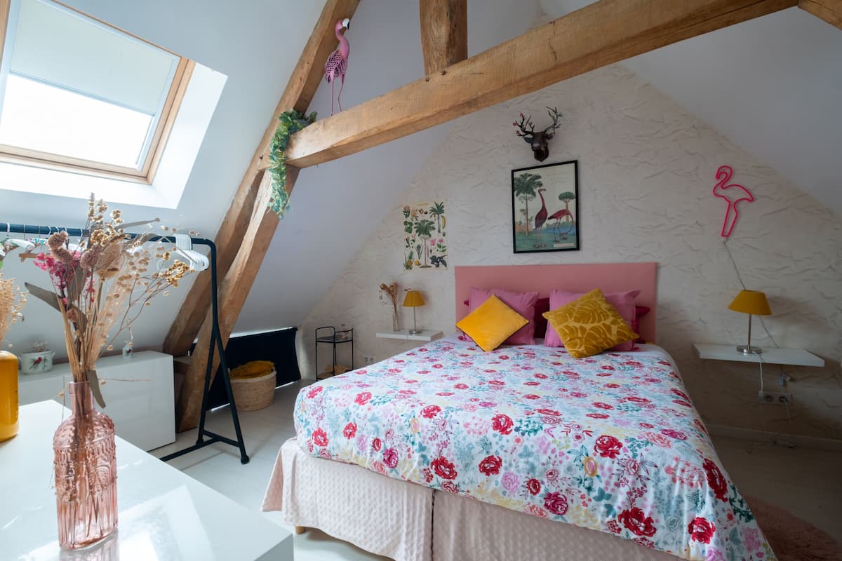 Un dormitorio con techo inclinado, una cama con ropa de cama floral, estructura de vigas de madera, un adorno de cabeza de venado colgado en la pared y una ventana con vistas al cielo.