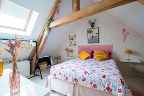 A bedroom with a sloped ceiling, a bed with floral bedding, a wooden beam structure, a framed picture, and a vase with dried flowers.