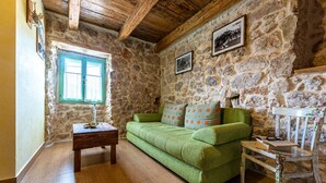 Una habitación con pared de piedra, vigas de madera en el techo, un sofá verde, una pequeña mesa de madera y una silla con estampado floral.