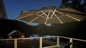 A deck with a table and chairs, an umbrella, and a view of a cabin and trees. - Downstairs Studio style apartment on the River (Mermentau)