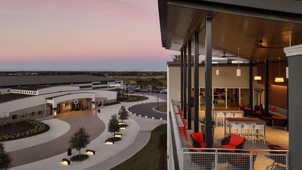 A hotel with a modern architectural design, featuring a spacious outdoor seating area with tables and chairs, a curved driveway, and a parking lot. - Residence Inn by Marriott Tampa Wesley Chapel (Wesley Chapel)