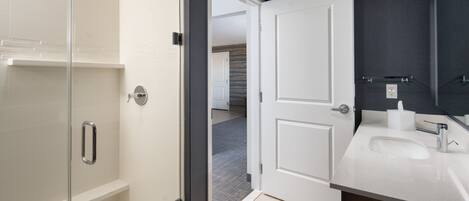 A modern bathroom with a glass shower enclosure, a white sink with a dark cabinet, and a door leading to another room.