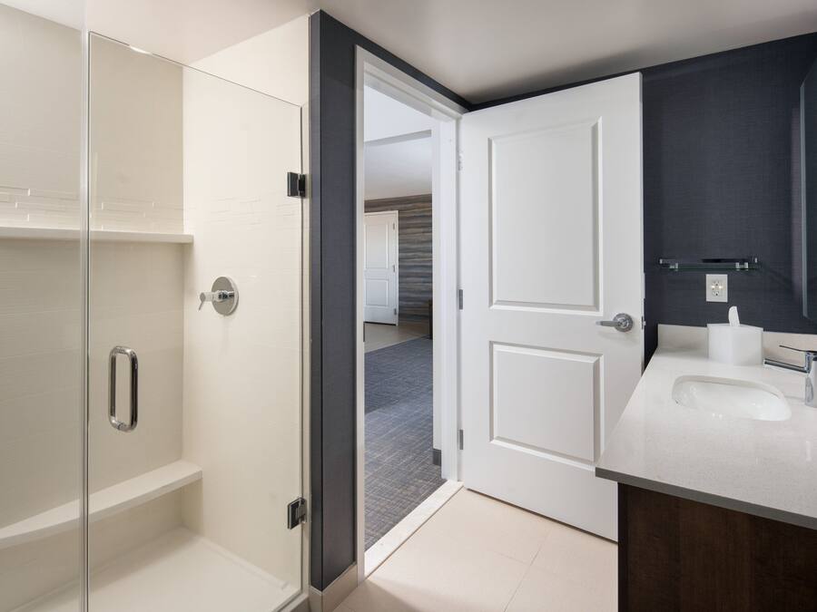 Een moderne badkamer met een glazen douchecabine, een wit wastafelmeubel met een donkere kast, en een deur die leidt naar een andere kamer.