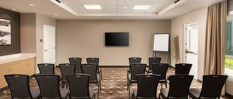 A conference room with a patterned carpet, black chairs, a flat-screen TV, a whiteboard, and a window with curtains.
