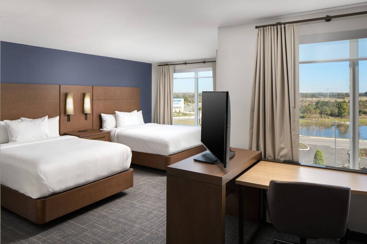 Ein Hotelzimmer mit zwei Betten, einem Schreibtisch, einem Sessel, einem Fernseher und einem großen Fenster mit Blick auf ein Gewässer und Grünflächen.