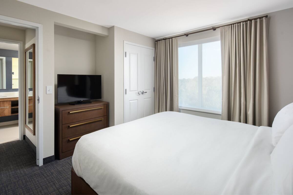 Ein Hotelzimmer mit Bett, Fernseher auf einem Nachttisch, einer Tür und einem mit Vorhängen versehenen Fenster.