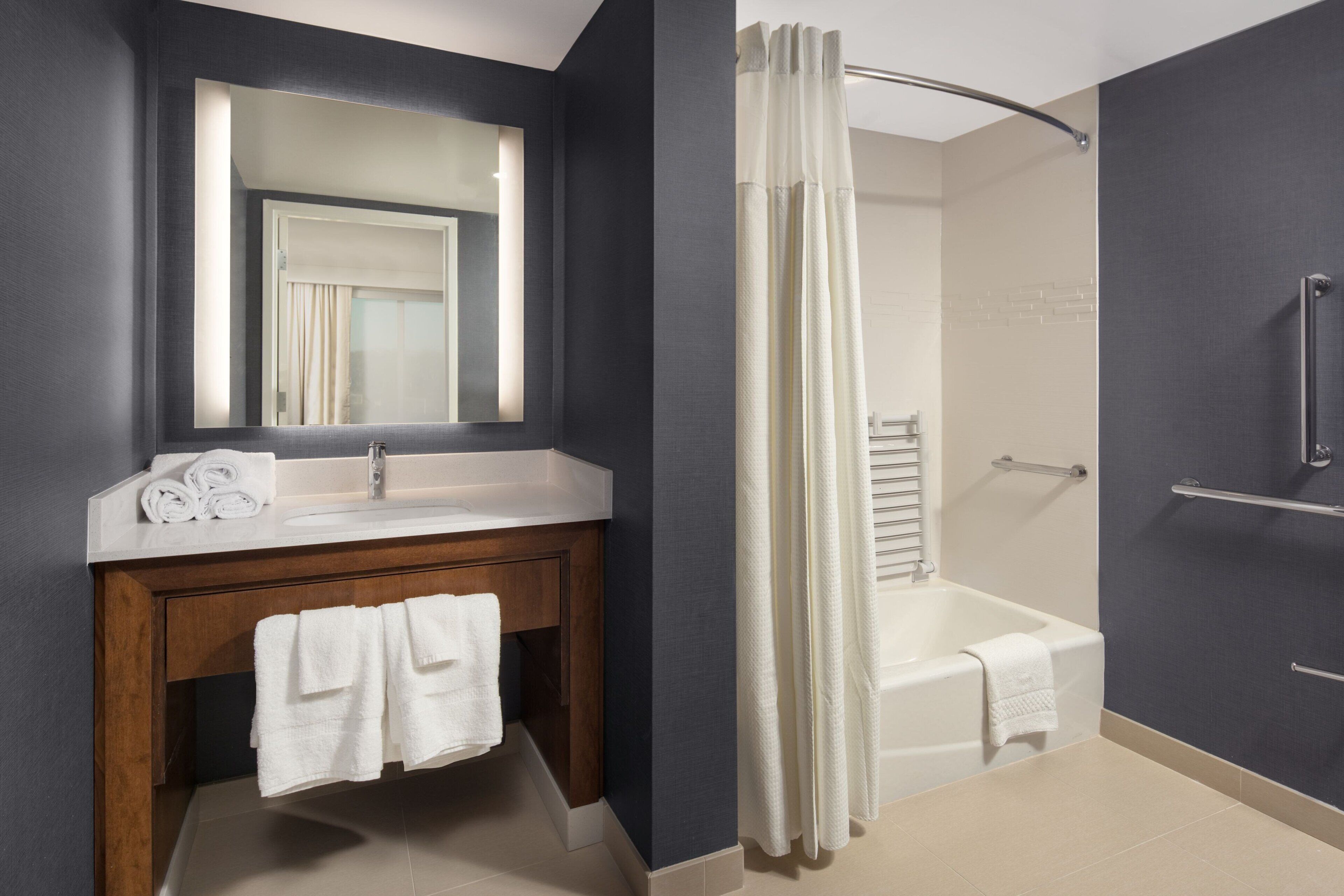 A modern bathroom with a dark wall, a white sink, a wooden vanity, a large mirror, a shower with a white curtain, and a bathtub.