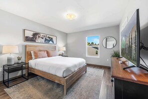 A modern bedroom with a wooden bed, a nightstand, a bedside table, a large window, a mirror, and a television. - Private Home, Full Kitchen, Laundry & Wi-Fi! (Wasilla)