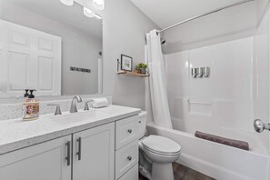 A modern bathroom with a white sink, vanity, and toilet. There is a shower curtain, a small shelf with a plant, and a hand soap dispenser. - Private Home, Full Kitchen, Laundry & Wi-Fi! (Wasilla)