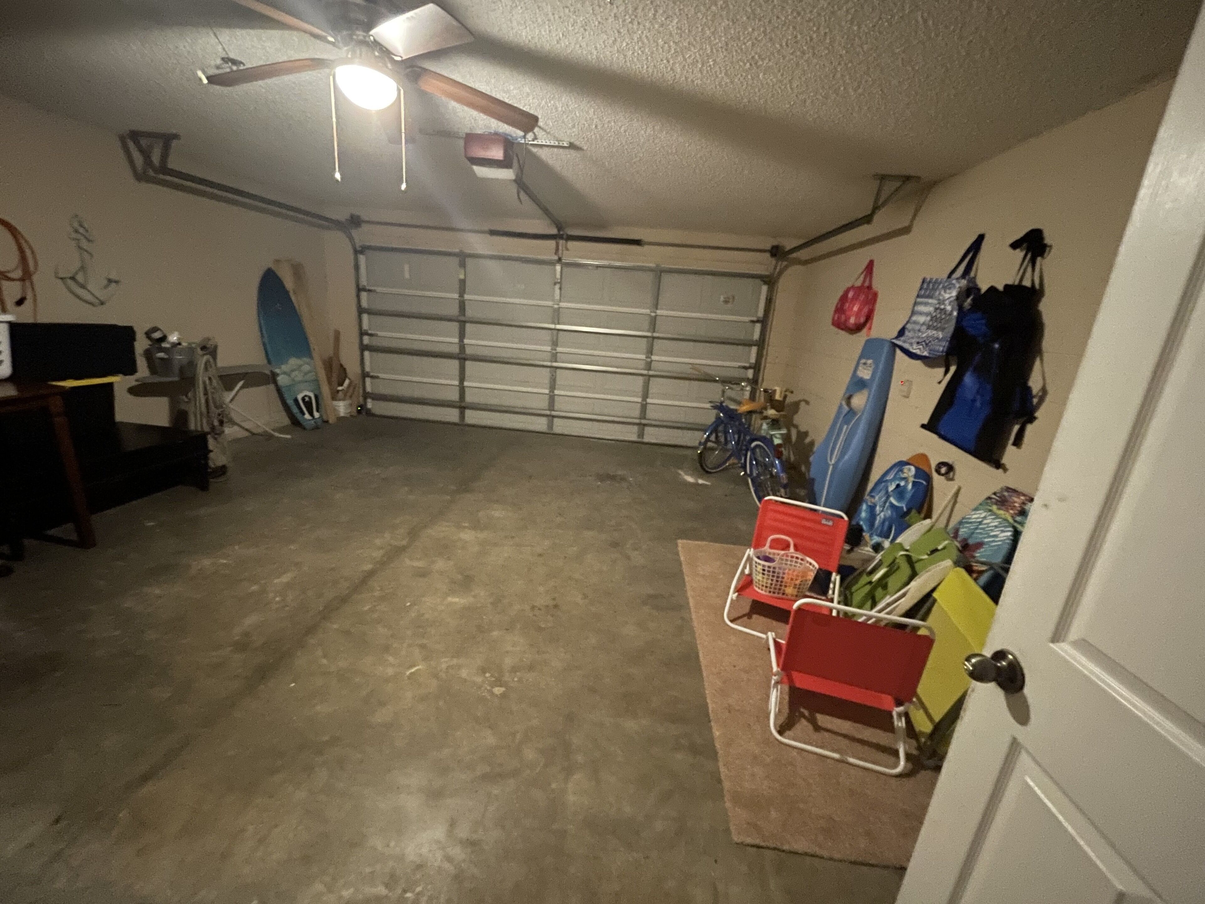 Un garage avec un sol en béton, un ventilateur de plafond, un vélo et une planche de surf appuyée contre le mur.