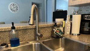 A kitchen with stainless steel sinks, a soap dispenser, a bottle of blue liquid soap, a paper towel holder, and a coffee maker.