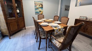 A dining area with a wooden table set for six, surrounded by six chairs with cushioned seats. A large painting of a bridge and sailboats hangs on the wall above the table.