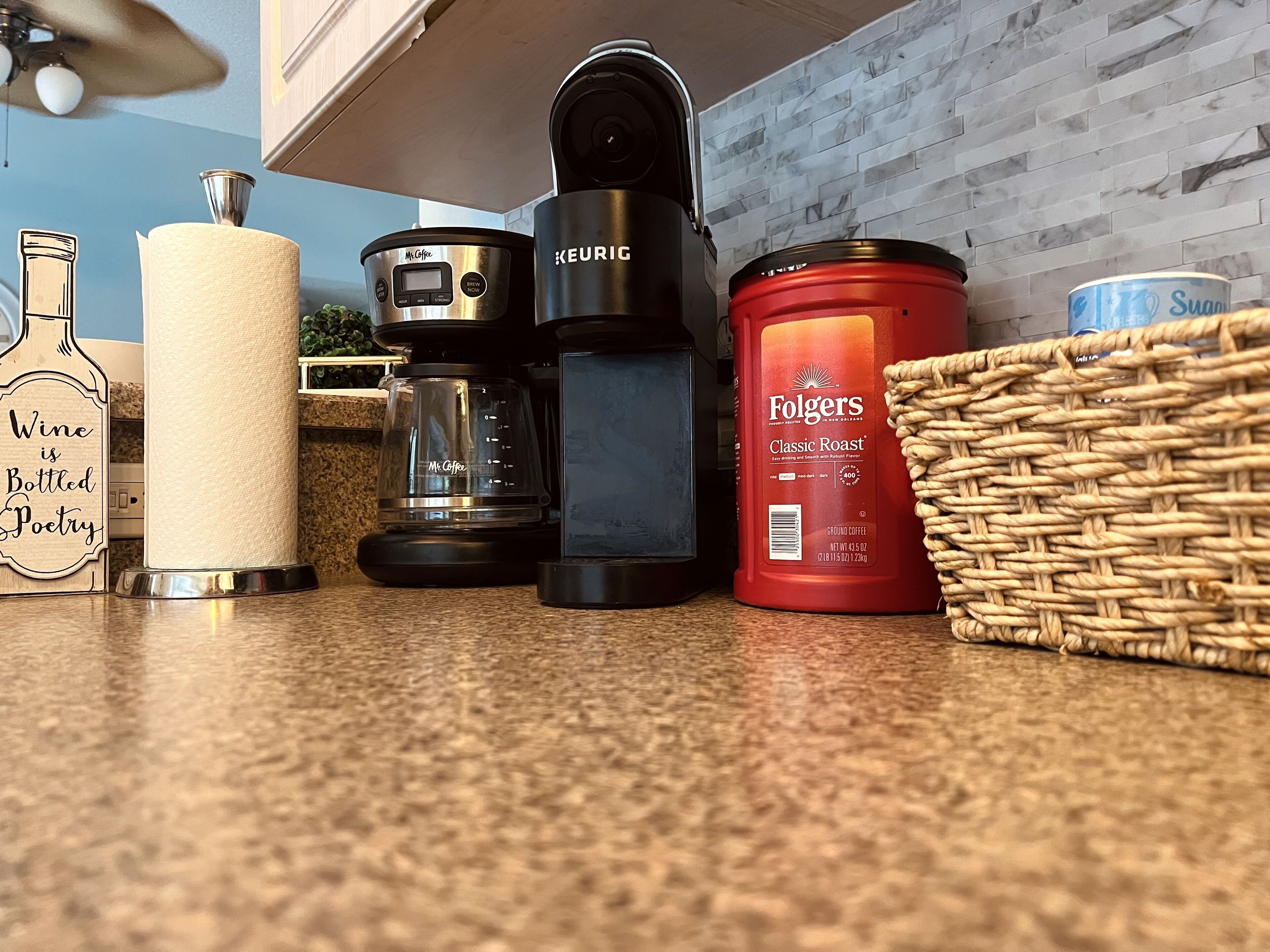 Een keukentafel met een koffiezetapparaat, een keukenrolhouder, een wijnfles en een mand.