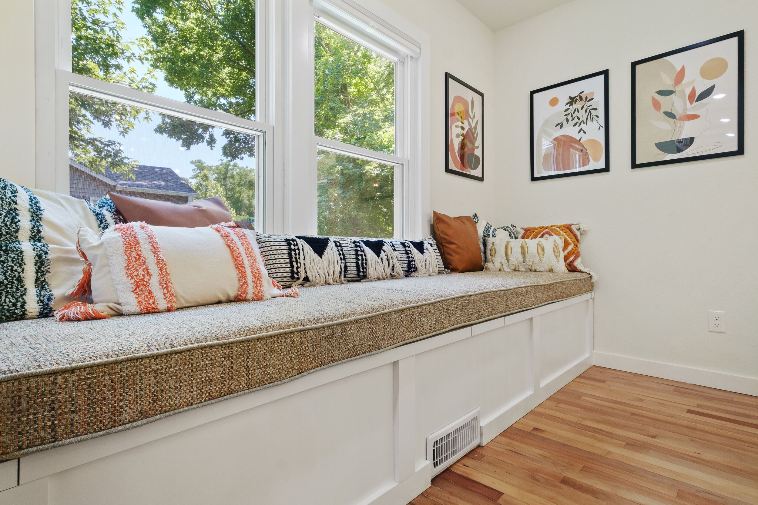 A room with a built-in bench along a window, white walls, and framed artwork on the wall.